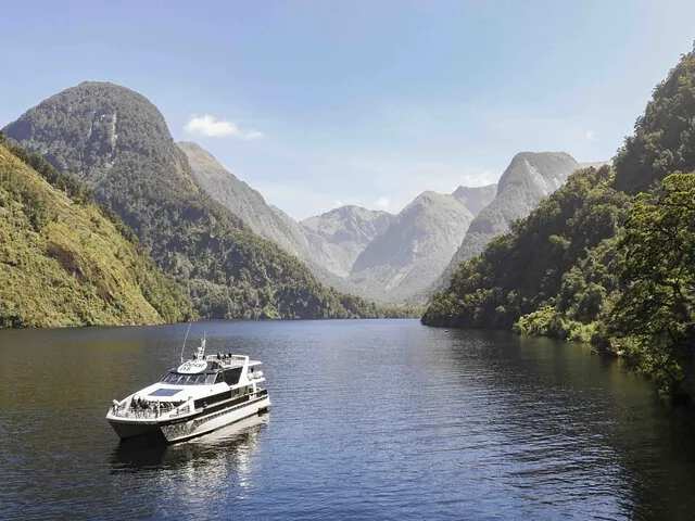 Ein weißes Ausflugsschiff fährt auf einem breiten Fluss zwischen grünen Bergen im Doubtful Sound.