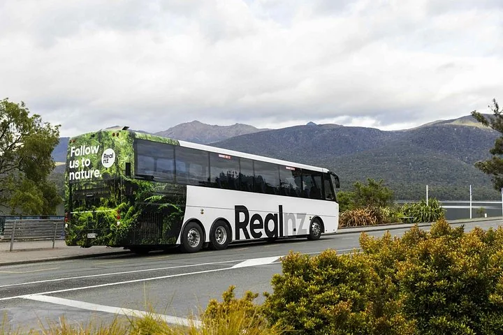 Ein weißer RealNZ-Tourbus fährt auf einer Straße mit Bergen im Hintergrund.
