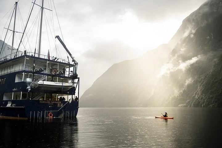 Ein einzelner Kajakfahrer neben einem großen Kreuzfahrtschiff, das im nebligen Doubtful Sound vor Anker liegt.